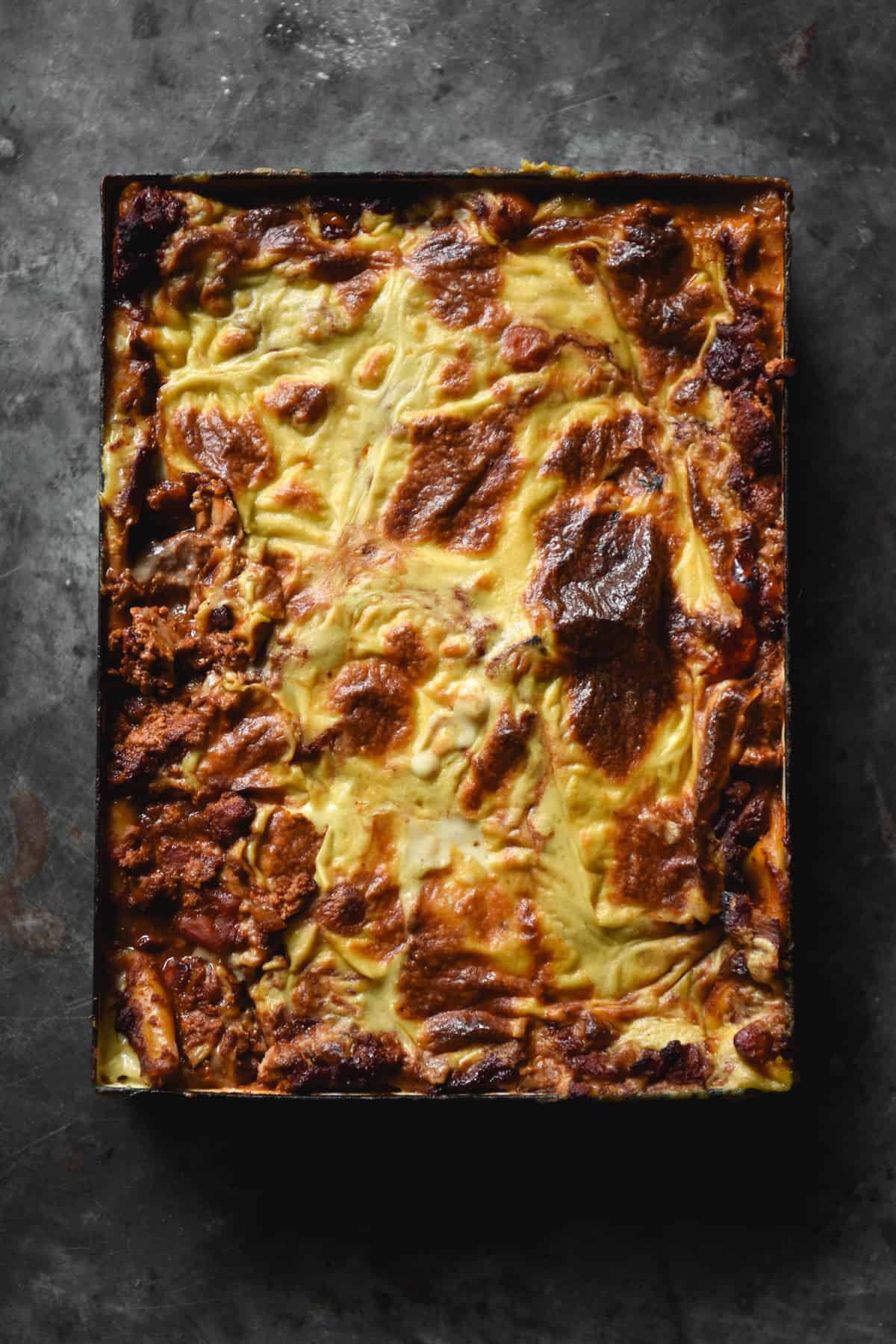 An aerial image of a rectangular baking dish filled with a golden brown vegan, gluten free and Low FODMAP lasagne. The lasagne sits atop a dark steel backdrop.