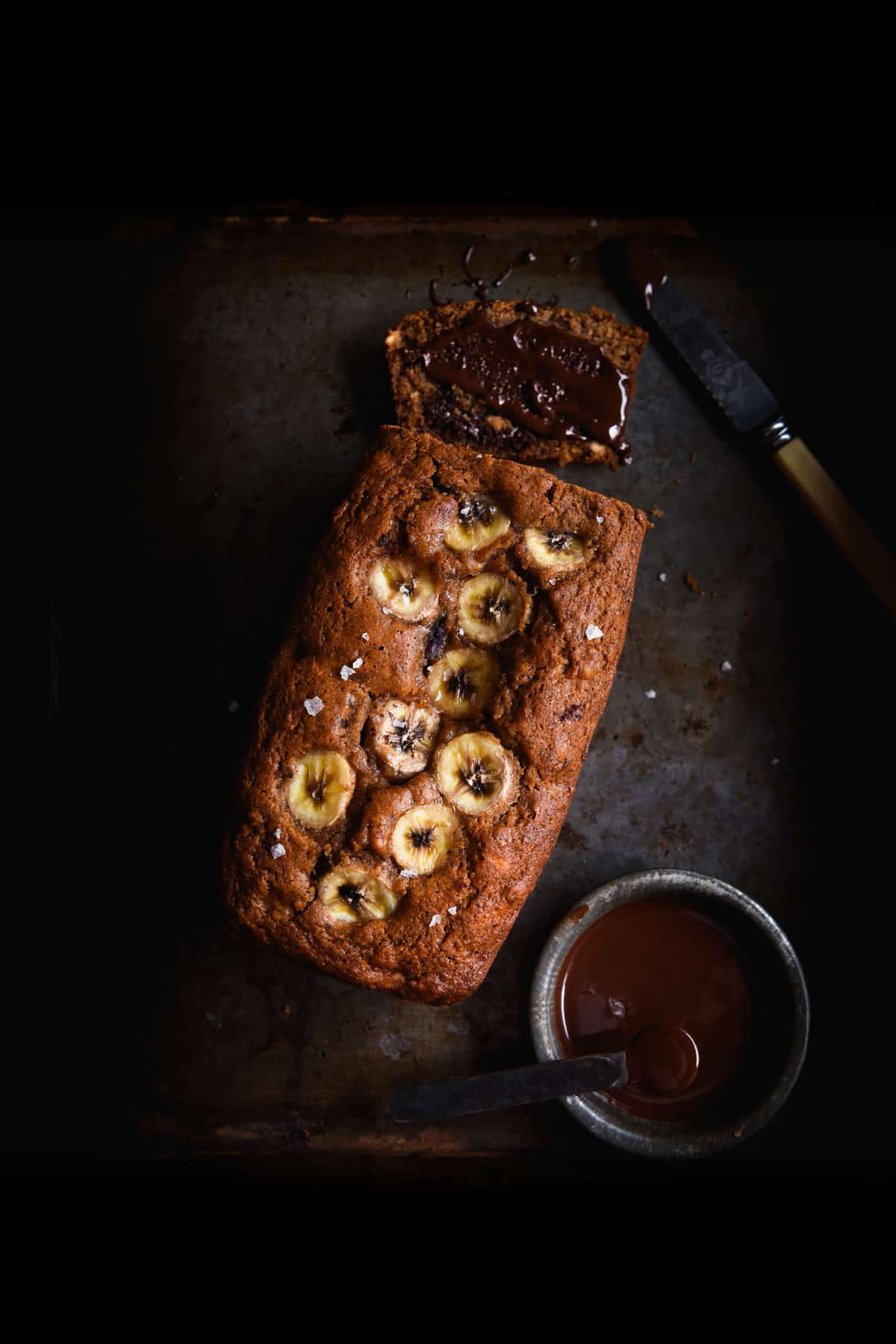 A moody aerial image of a loaf of gluten free banana bread on a dark steel backdrop. The loaf is golden brown and topped with banana coins. A slice of the bread has been smeared with chocolate spread, a bowl of which sits to the bottom right of the image.