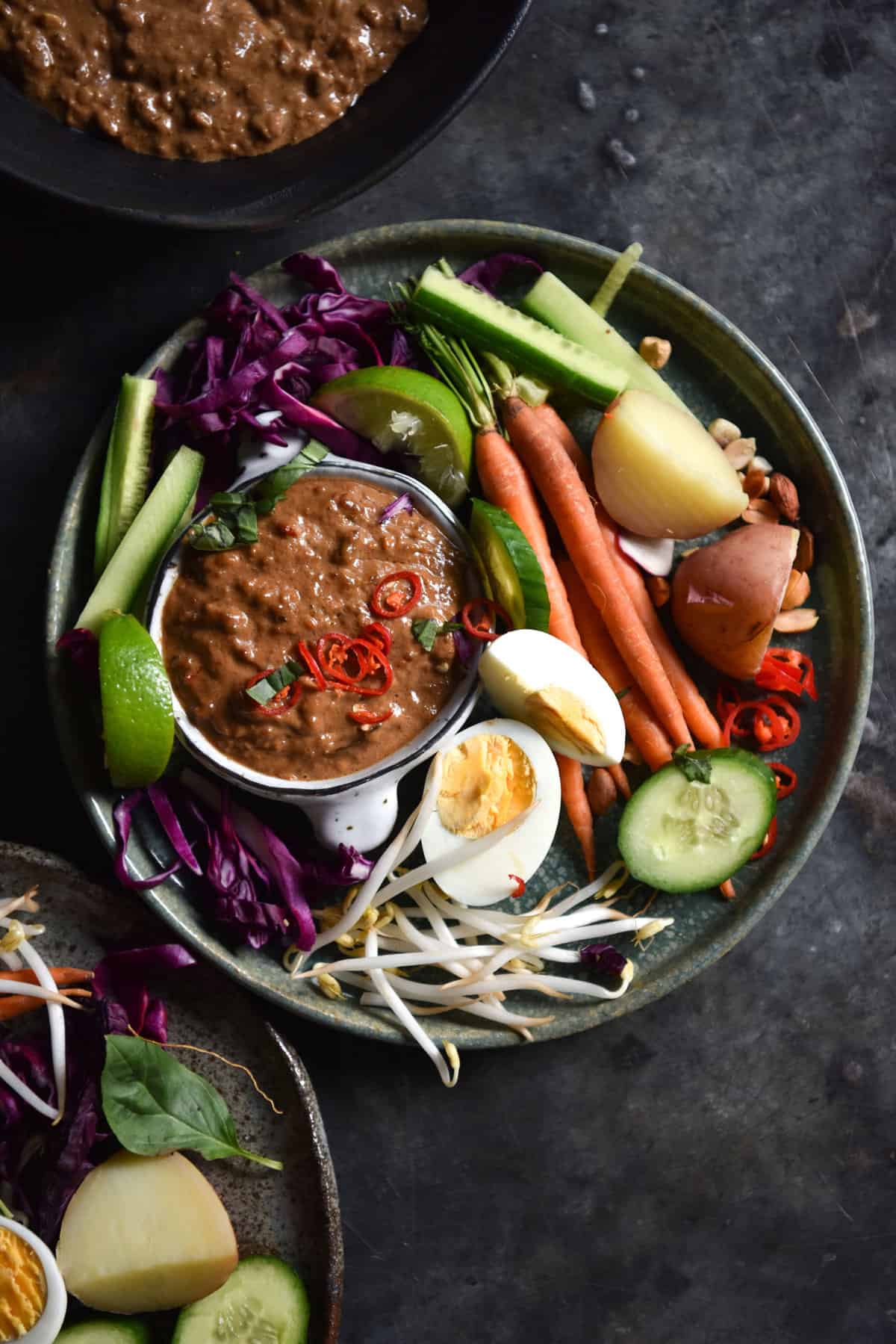 Low FODMAP peanut satay sauce + a quick gado gado on a steel dark blue backdrop. The satay is in a central bowl on the plate, and the gado gado vegetables are arranged around the outside.