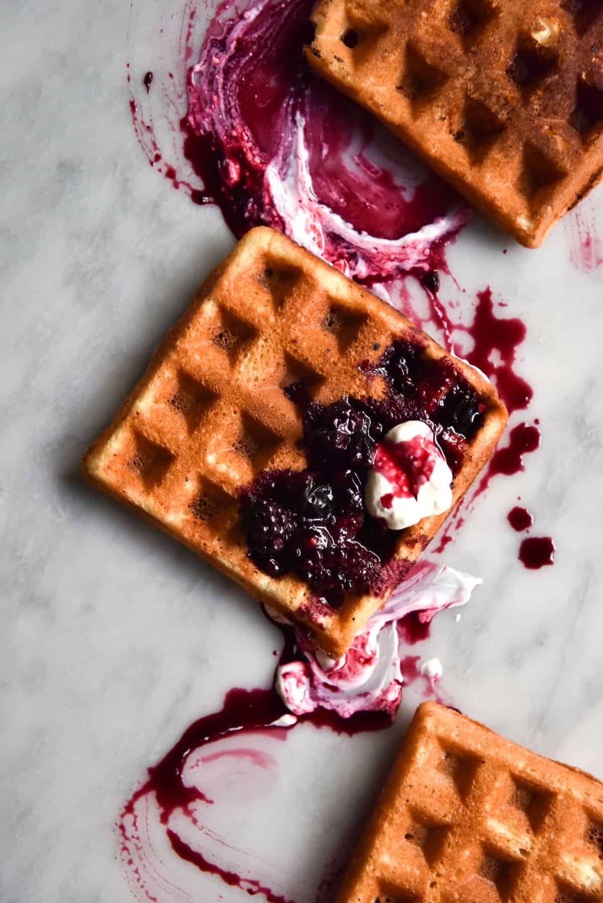 An aerial image of gluten free waffles atop a white marble table. The waffles are golden brown and topped with yoghurt and a berry compote which creates a beautiful marbled pattern over the waffle and the table.