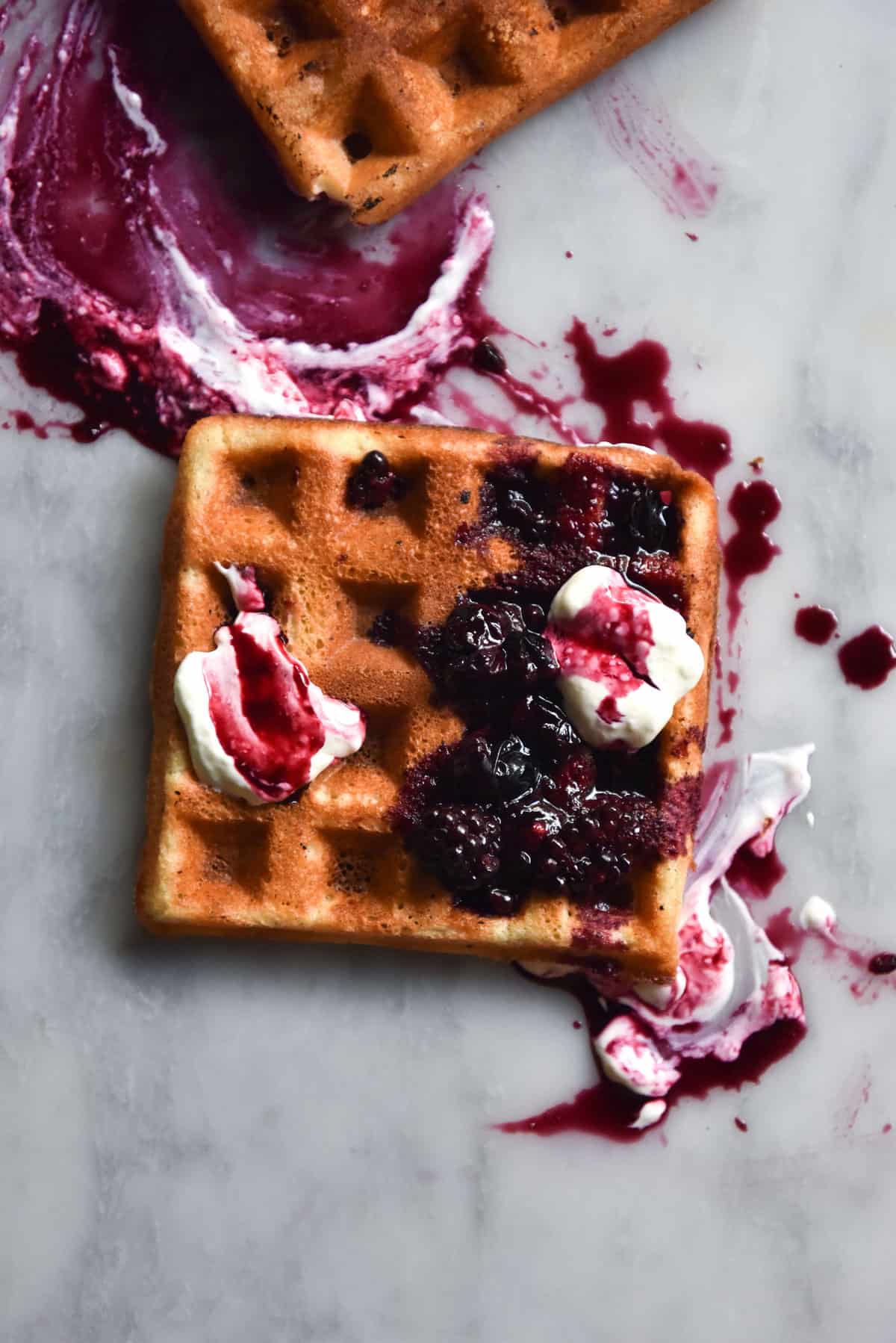 An aerial image of a gluten free waffle atop a white marble table. The waffle is golden brown and topped with yoghurt and a berry compote which creates a beautiful marbled pattern over the waffle and the table.