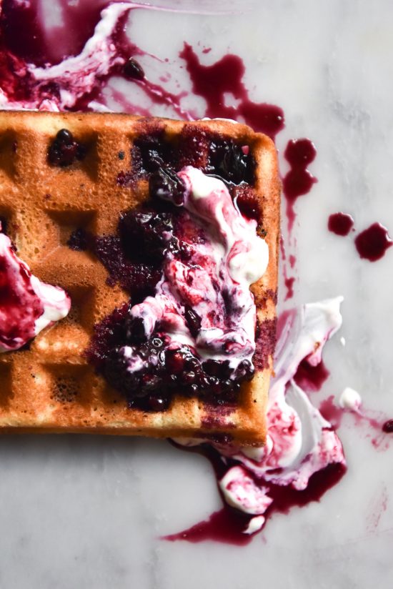 An aerial close up image of a gluten free waffle topped with swirled yoghurt and berry compote on a white marble backdrop
