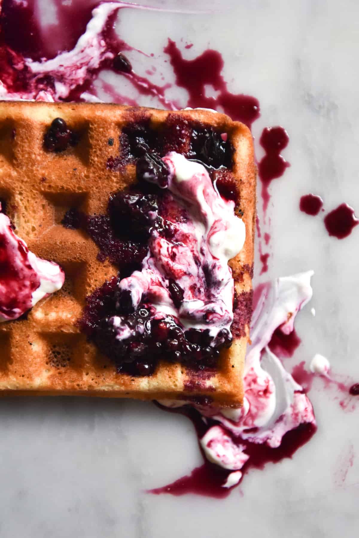 An aerial image of a gluten free waffle atop a white marble table. The waffle is golden brown and topped with yoghurt and a berry compote which creates a beautiful marbled pattern over the waffle and the table.