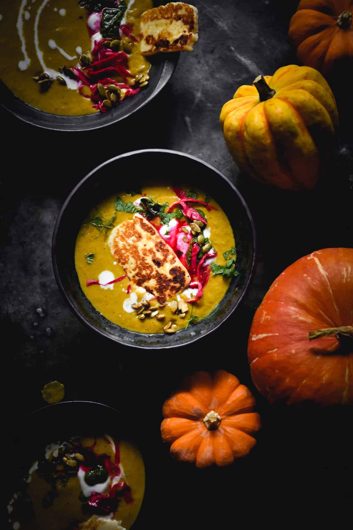 A moody aerial image of a dark ceramic bowl filled with Moroccan spiced pumpkin soup. The bowl is topped with haloumi, herbs, pepitas and pickled cabbage. The central bowl is surrounded by heirloom pumpkins.