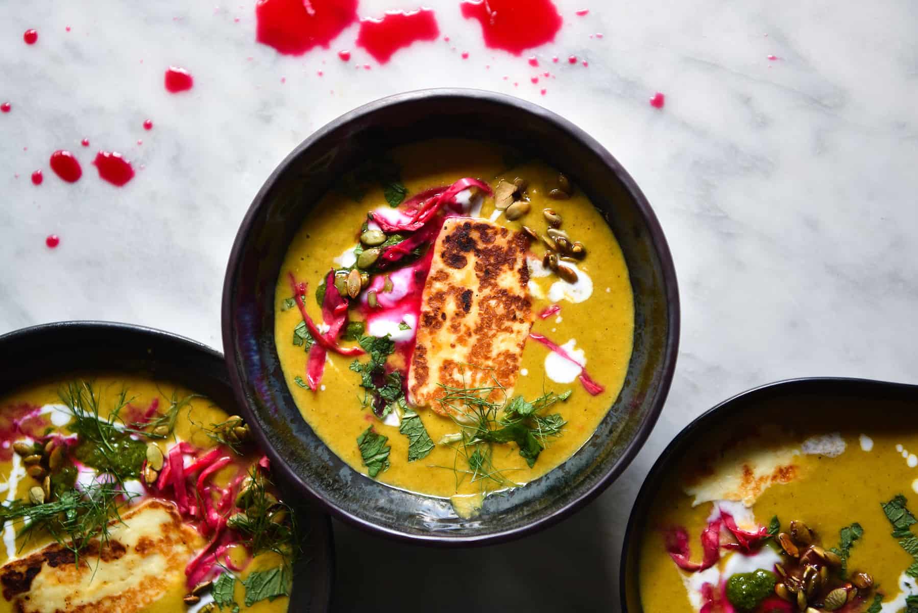An aerial image of three dark ceramic bowl filled with Moroccan spiced pumpkin soup atop a white marble table. The bowls are topped with haloumi, herbs, pepitas and pickled cabbage. 