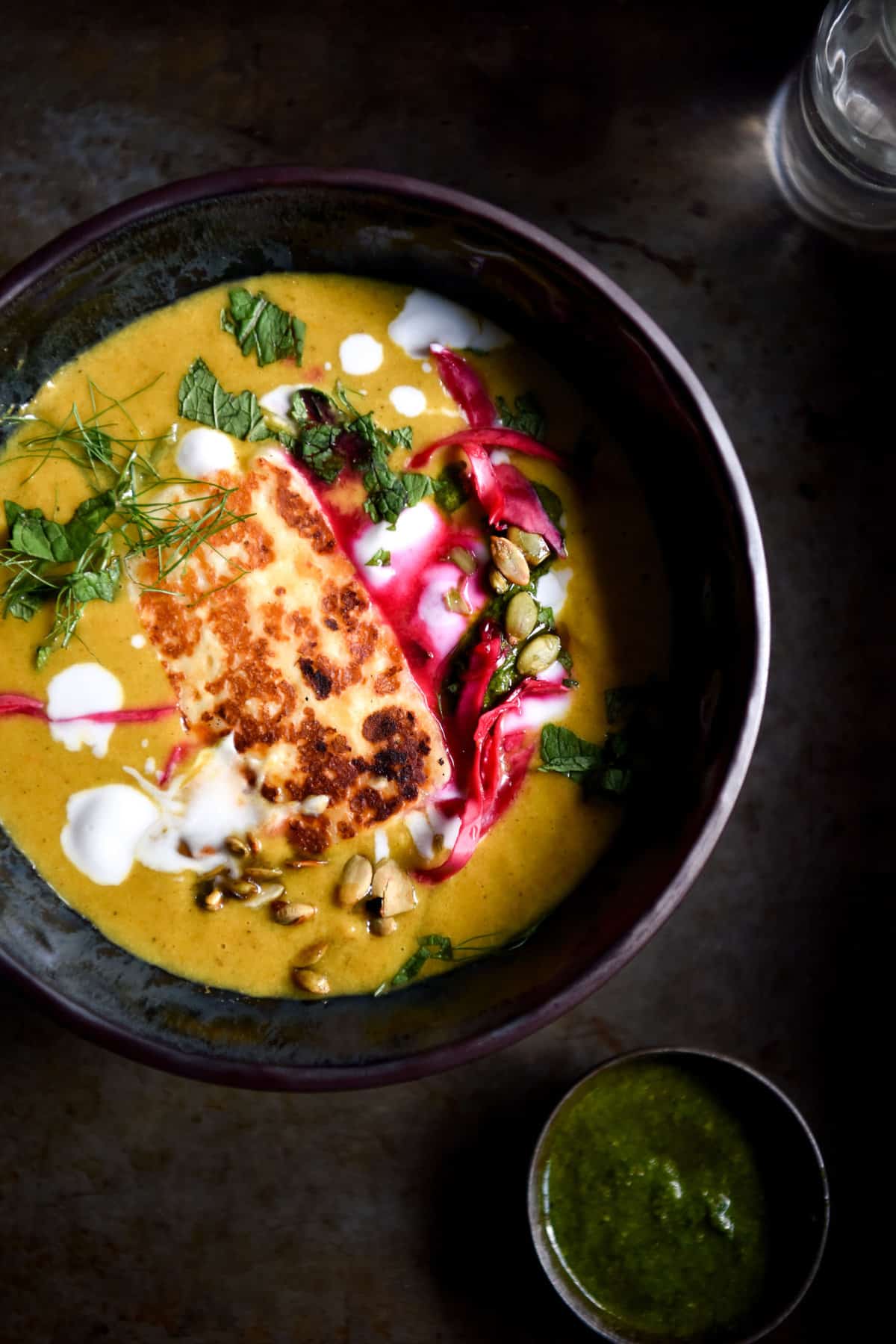 Moroccan spiced pumpkin soup in a dark grey ceramic bowl atop a dark grey metal backdrop. The soup is topped with a piece of fried haloumi, pickled cabbage, herbs, cream and pepitas.