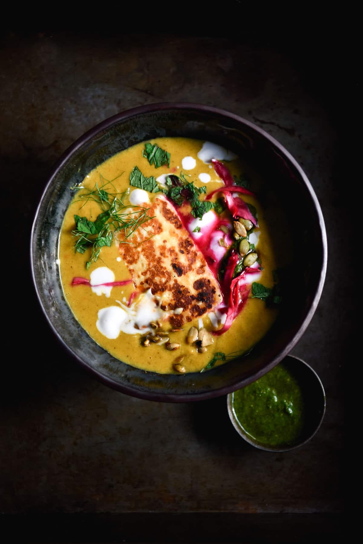 A moody aerial image of a dark ceramic bowl filled with Moroccan spiced pumpkin soup. The bowl is topped with haloumi, herbs, pepitas and pickled cabbage. 
