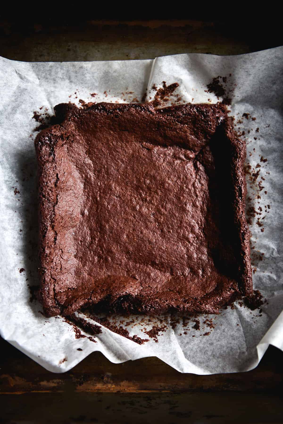 A moody aerial image of a slab of gluten free vegan brownie on a crinkled sheet of baking paper atop a dark steel backdrop