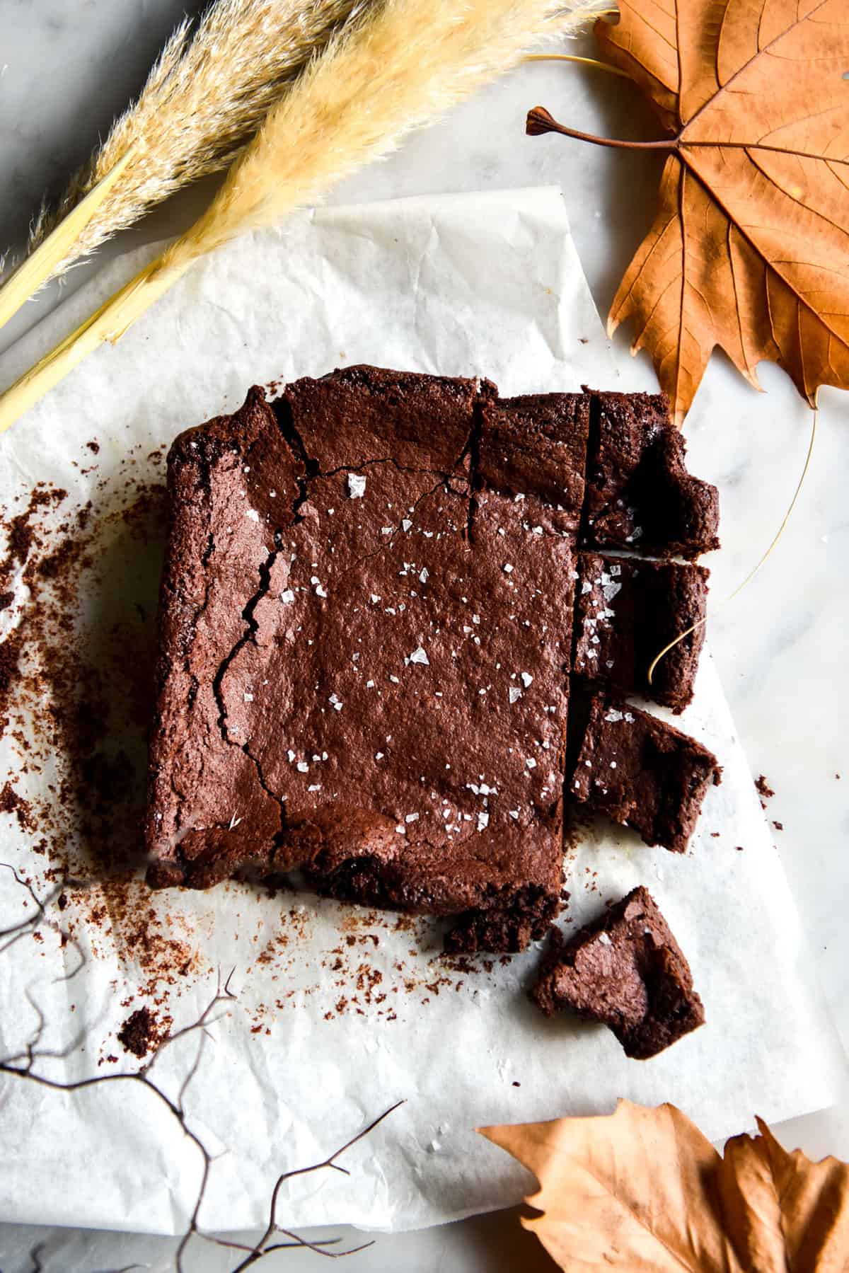 An aerial image of a slab of gluten free vegan brownie atop a white marble table. The brownie is sprinkled with sea salt flakes and the table is decorated with fall themed leaves and florals.