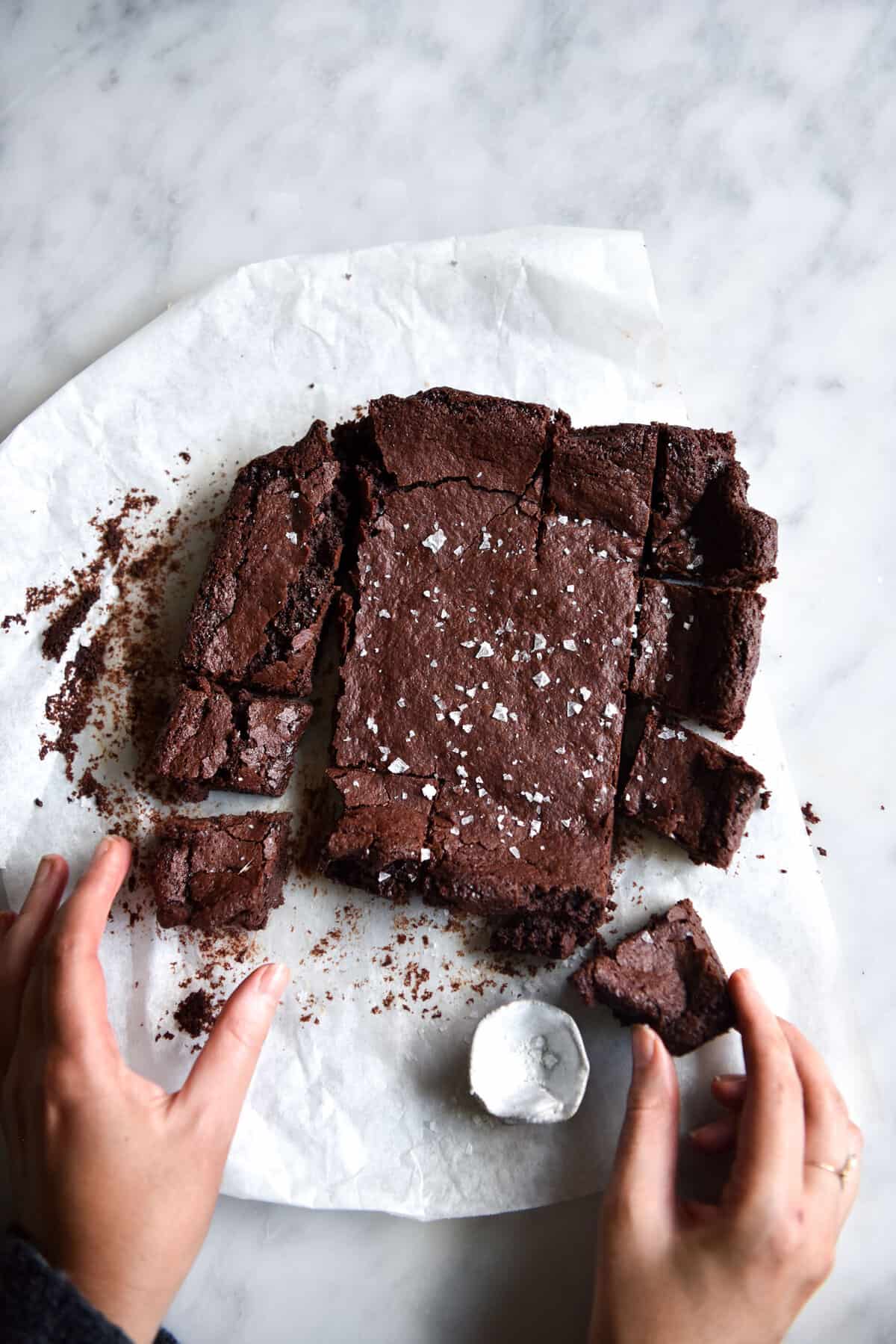 An image of a slab of gluten free vegan brownie atop a white marble table. The brownie is on a white sheet of baking paper and two hands extend from the bottom of the image to take pieces of the sliced brownie.
