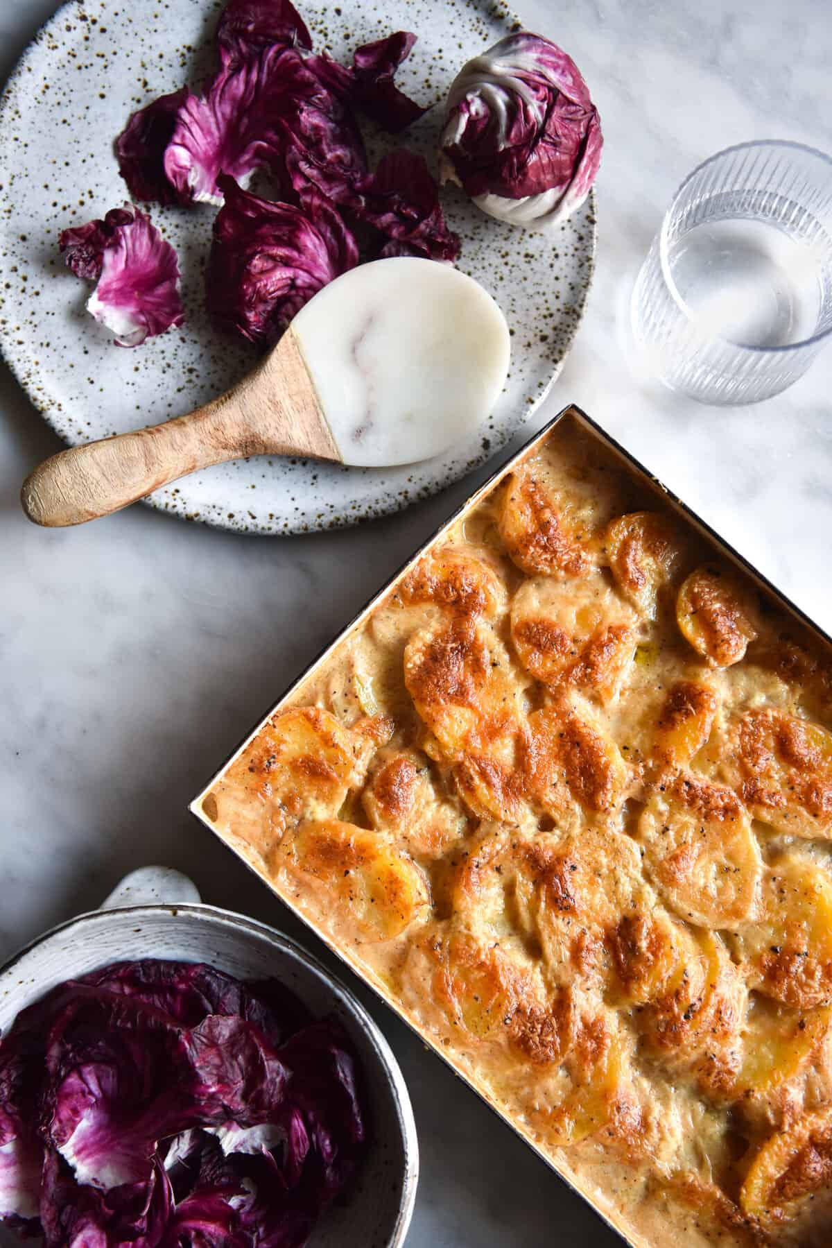 An aerial image of a tray of cacio e pepe potato bake on a white marble table surrounded by white plates topped with radicchio salad.
