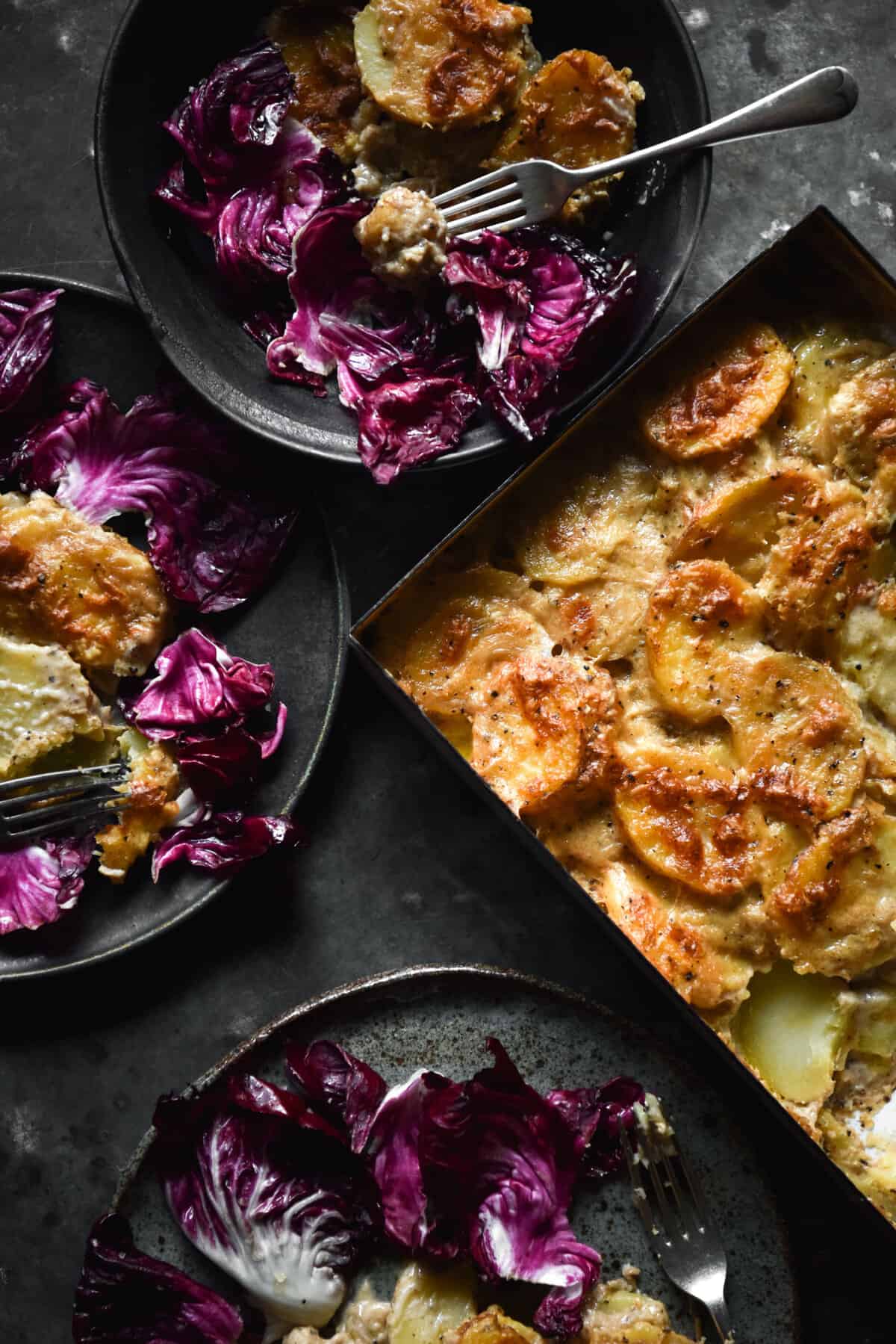 A moody aerial image of plates of cacio e pepe potato bake and radicchio salad on a dark blue steel backdrop. 