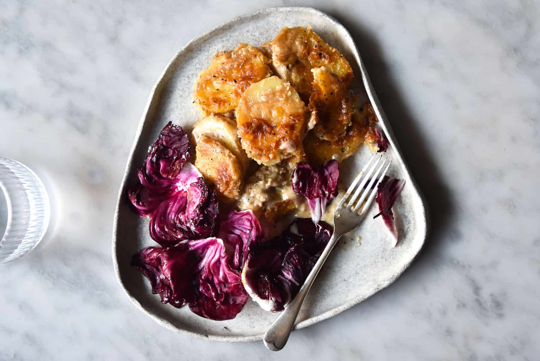 An aerial image of a white ceramic plate topped with radicchio salad and a cacio e pepe potato bake. The plate sits atop a white marble table and a glass of water sits to the left.