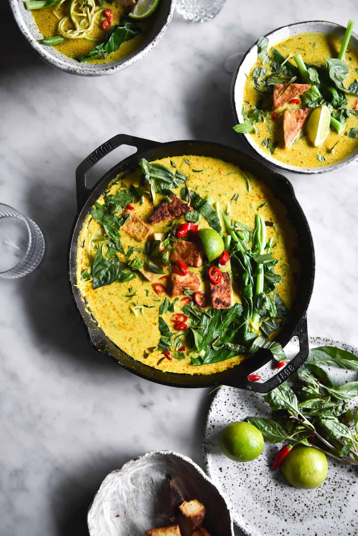 An aerial image of a vegan laksa in a black skillet atop a white marble table. The laksa has a golden yellow hue and is topped with fresh herbs, chilli, tofu puffs and a lime wedge.