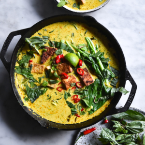 A skillet filled with FODMAP friendly vegan laksa topped with tofu cubes, chilli, lime wedges, greens and herbs. The skillet sits on a white marble table and is surrounded by other bowls of laksa.