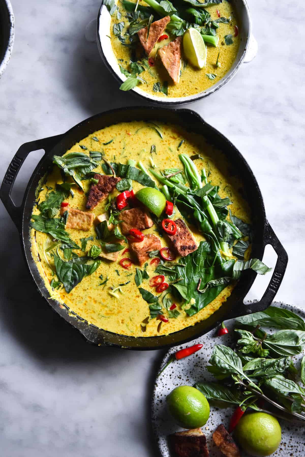 A skillet filled with FODMAP friendly vegan laksa topped with tofu cubes, chilli, lime wedges, greens and herbs. The skillet sits on a white marble table and is surrounded by other bowls of laksa.