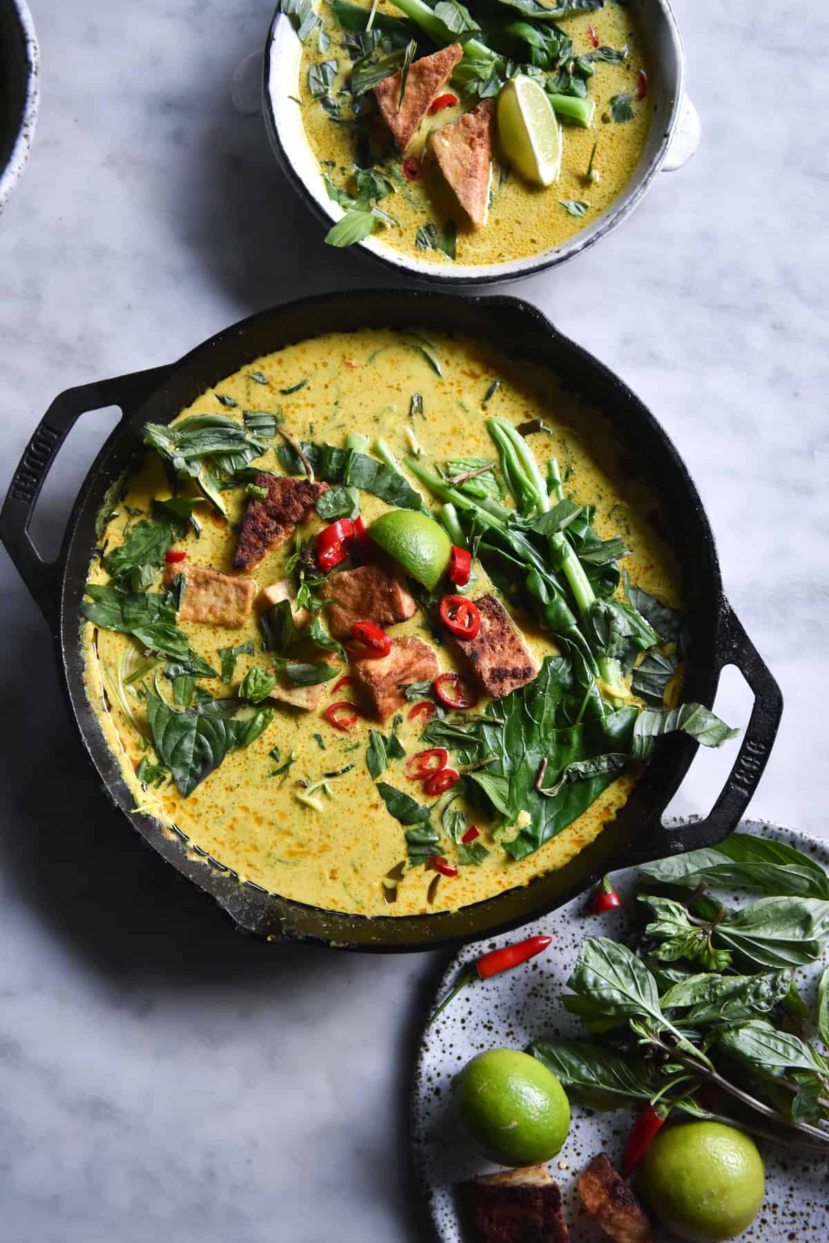 An aerial image of a vegan laksa in a black skillet atop a white marble table. The laksa has a golden yellow hue and is topped with fresh herbs, chilli, tofu puffs and a lime wedge.
