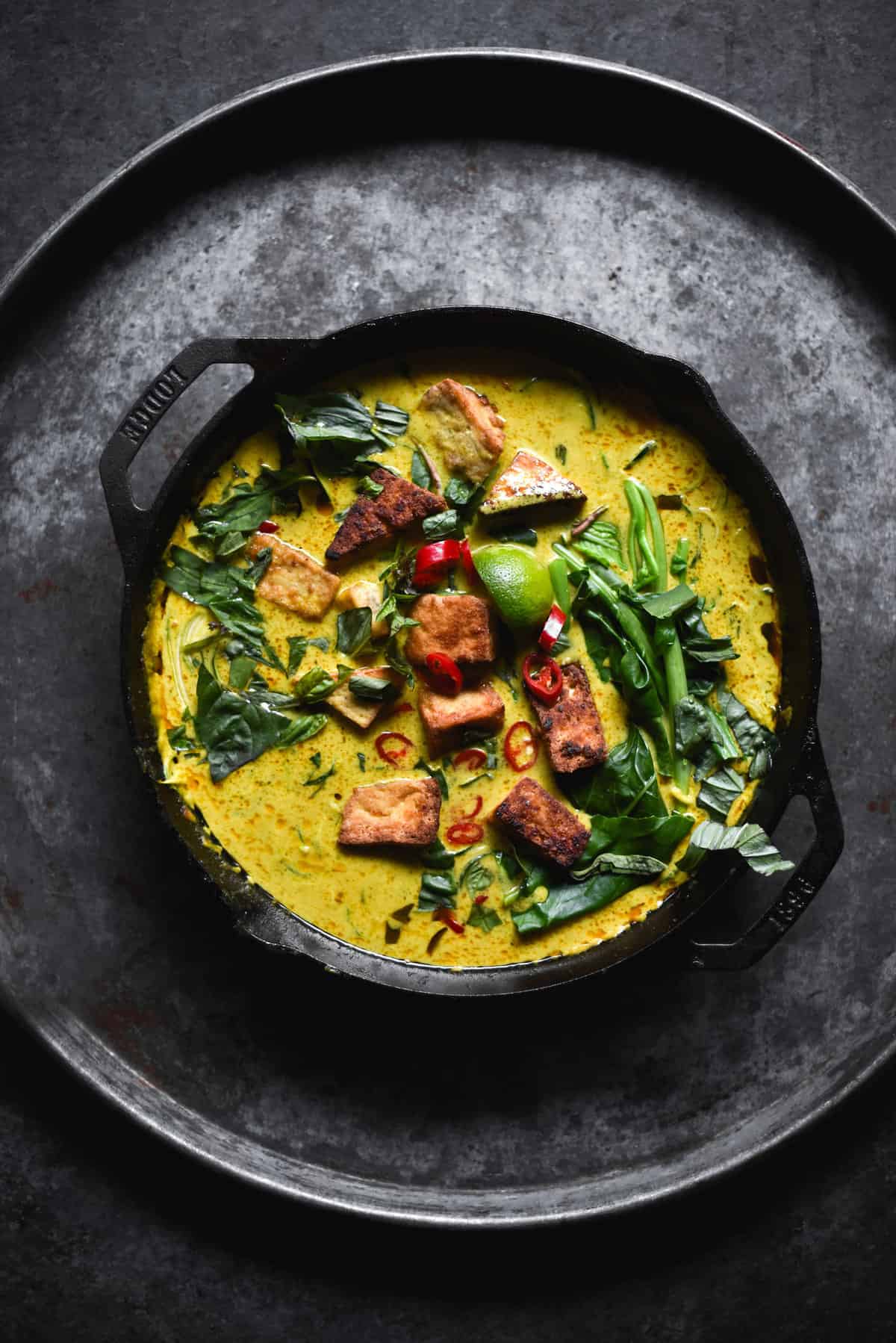 A skillet filled with FODMAP friendly vegan laksa topped with tofu cubes, chilli, lime wedges, greens and herbs. 