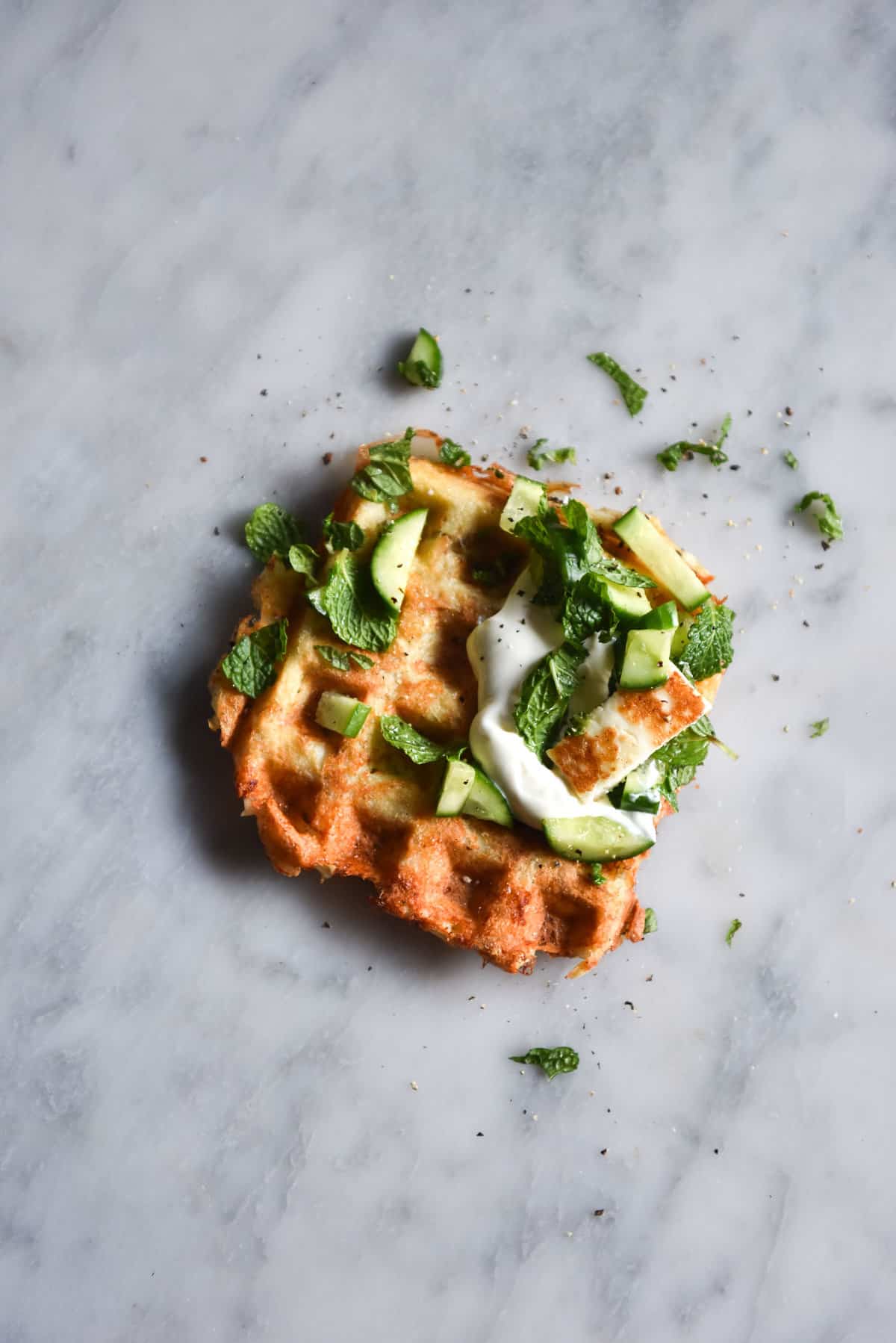 An aerial image of a three ingredient potato waffle atop a white marble table. The waffle is golden brown and topped with cucumbers, herbs, yoghurt and a slice of haloumi