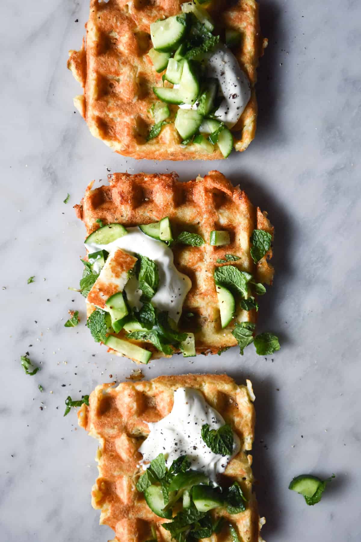 An aerial image of three potato waffles lined up on a white marble table. The waffles are topped with cucumbers, herbs, yoghurt and small pieces of haloumi.