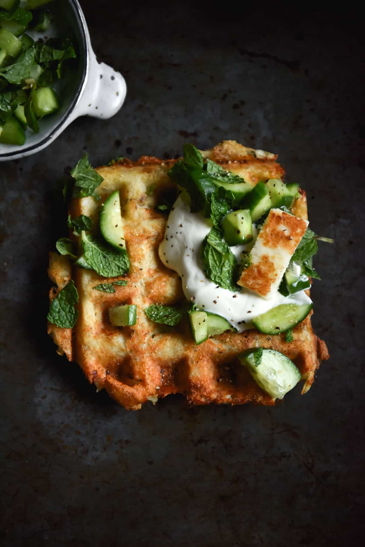 An aerial image of a three ingredient potato waffle atop a dark and moody backdrop. The waffle is topped with cucumbers, herbs, yoghurt and a slice of haloumi.