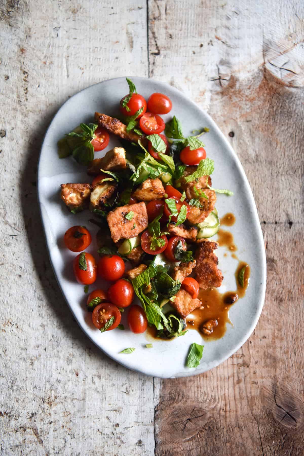 An aerial image of a salt and pepper tofu salad with tomatoes and herbs on a white plate atop a wooden table. 