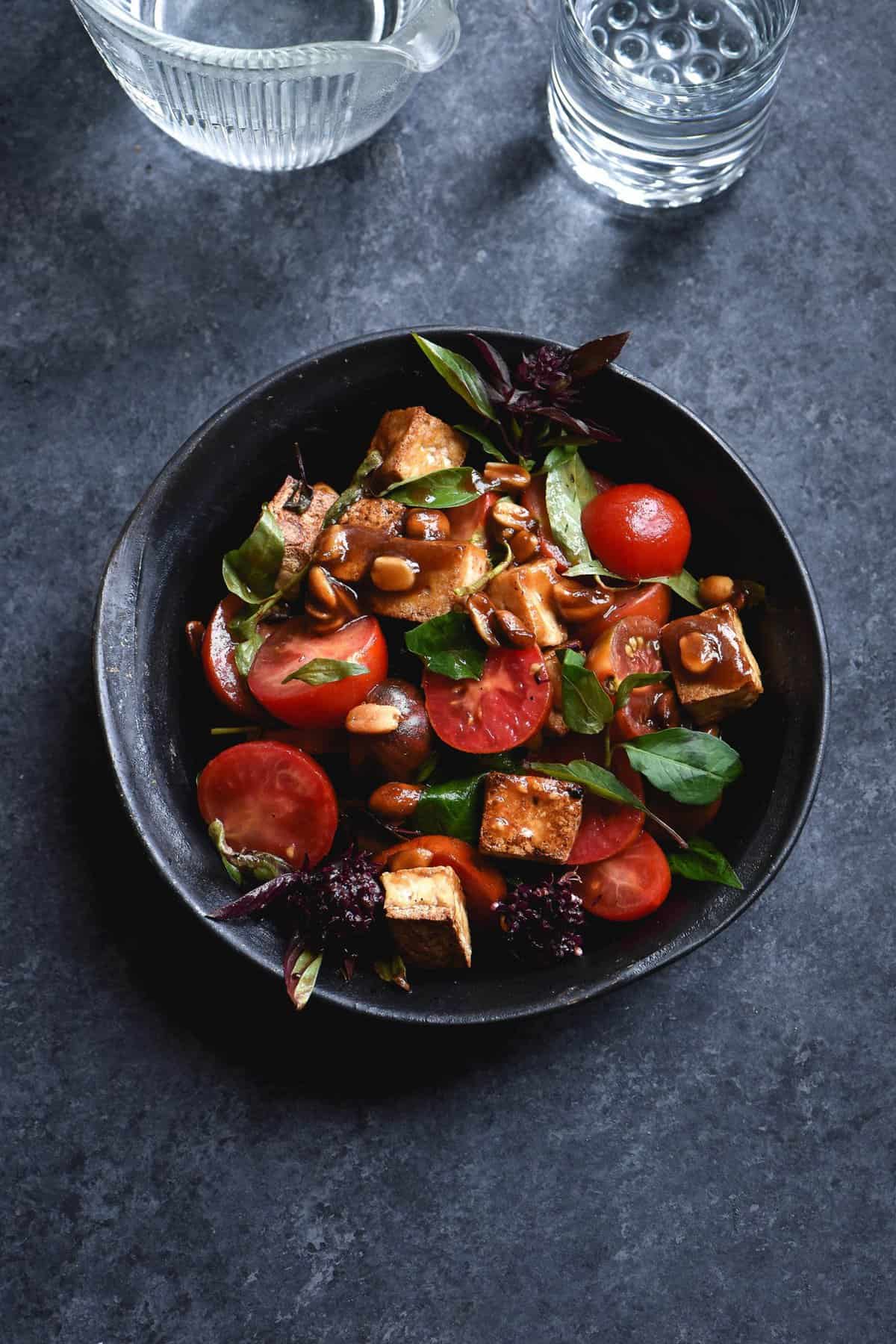 Salt and pepper tofu salad with tomatoes, toasted peanuts and a sweet tamarind makrut lime dressing against a dark blue backdrop with water glasses in the background