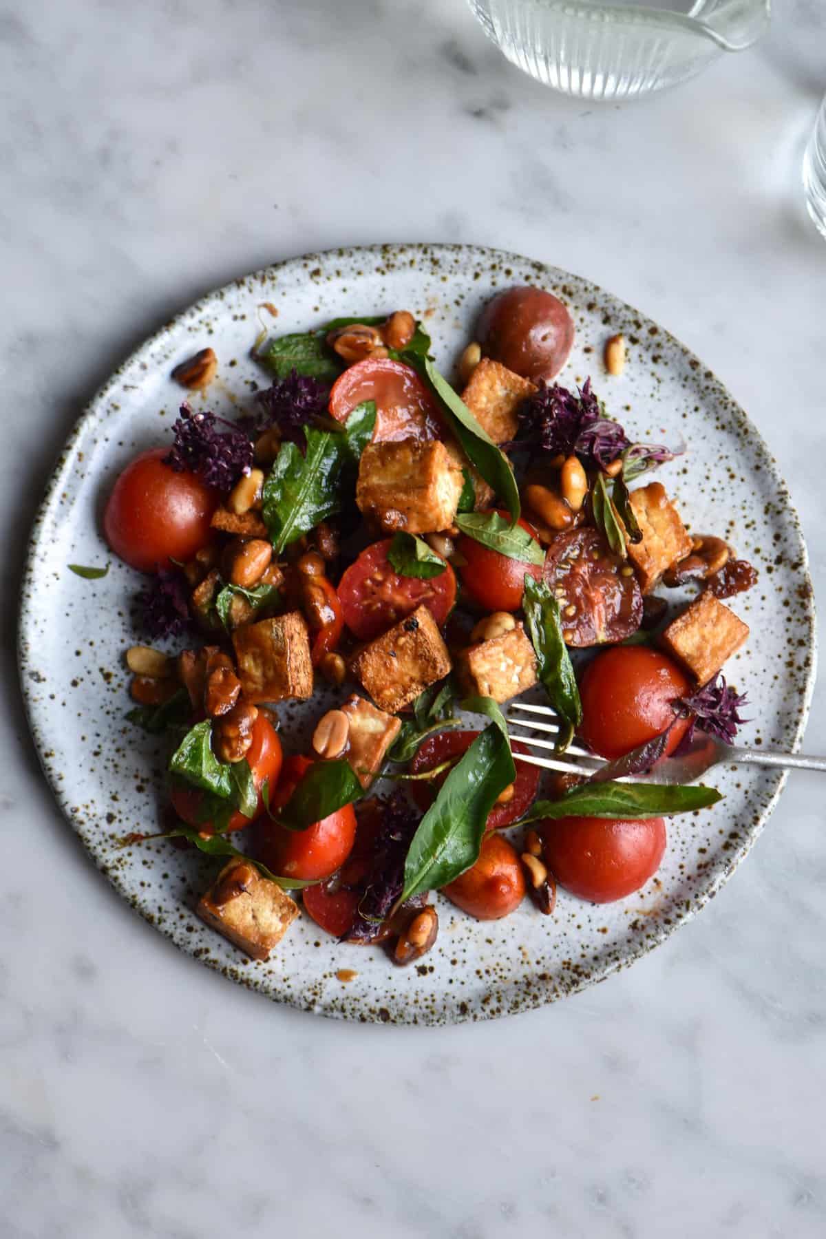 A salt and pepper tofu salad with tomatoes, herbs and a tamarind makrut lime dressing. Plated on a white ceramic plate against a white marble table