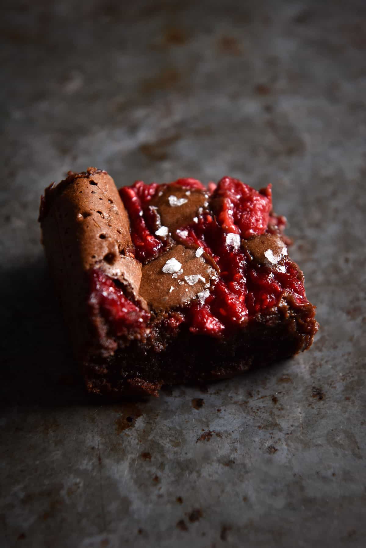 A moody side on image of a gluten free brownie topped with raspberries and sea salt flakes atop a dark steel backdrop