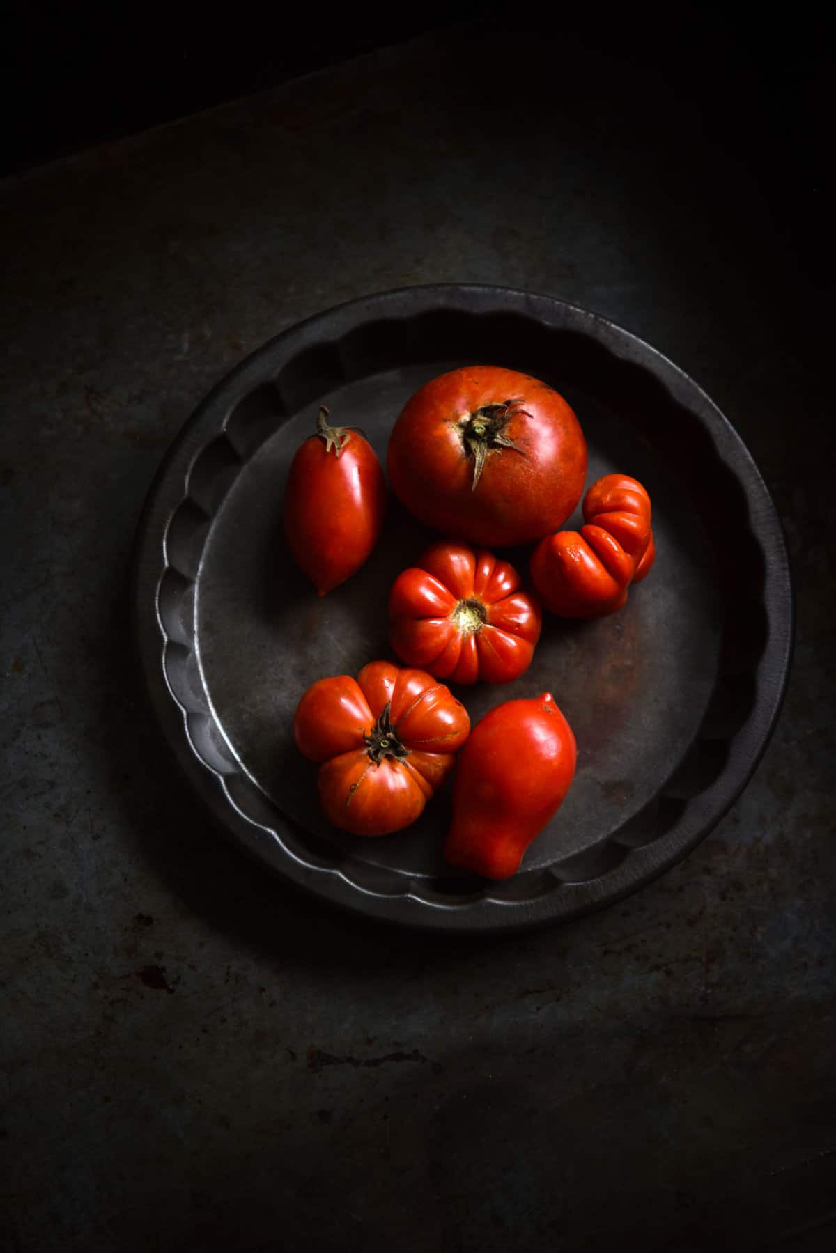 A macro moody image of heirloom tomatoes in a vintage steel baking tin atop a dark backdrop