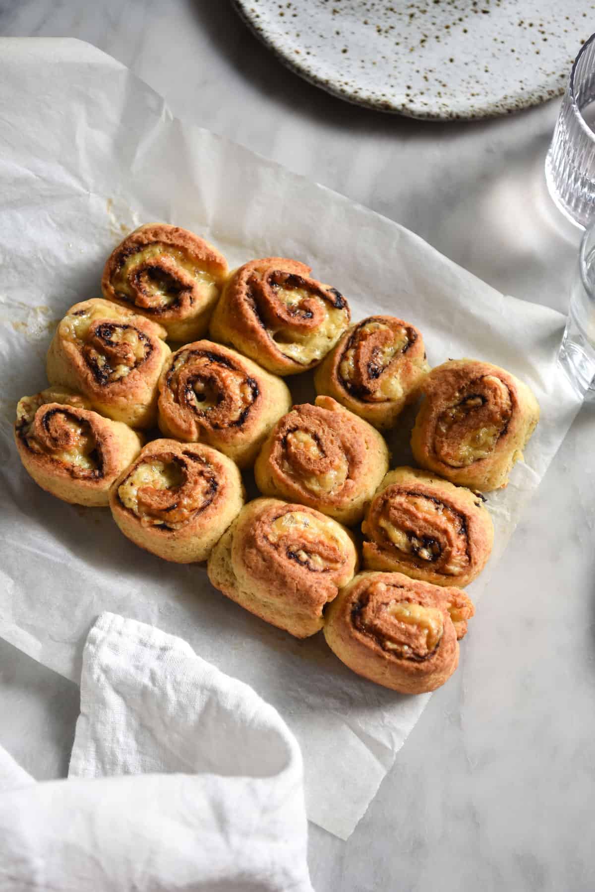 An aerial image of gluten free Vegemite scrolls on a white marble table. The scrolls are grouped together and surrounded by a white plate, water glasses and a linen tablecloth.