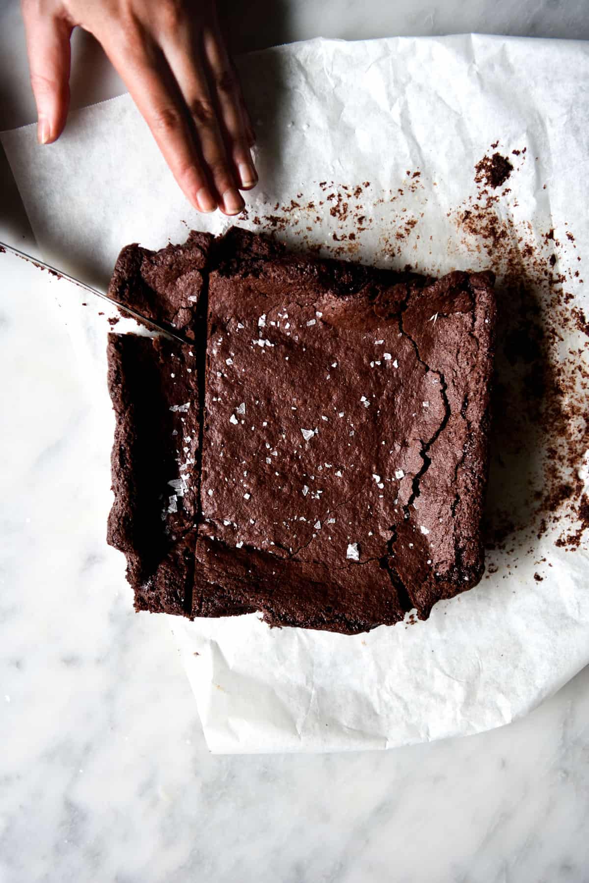 An aerial image of a slab of gluten free vegan brownies on a white marble table. A hand extends out from the top left of the image to slice into the brownie square.