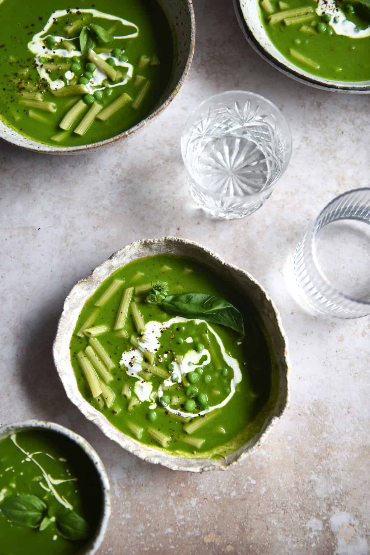 An aerial image of three white ceramic bowls of low FODMAP pesto pasta soup on a beige backdrop surrounded by glasses of water