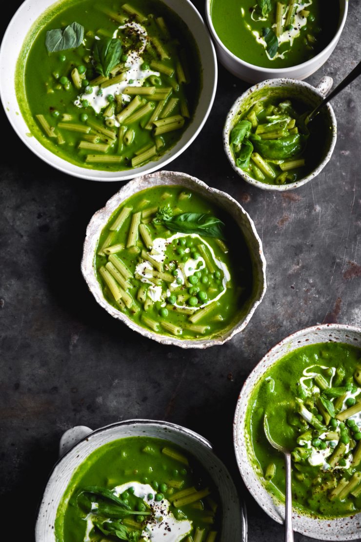 An aerial view of six white ceramic bowls filled with a bright green FODMAP friendly pesto pasta soup. The bowls are topped with a swirl of cream some extra basil leaves. They sit atop a dark blue steel backdrop