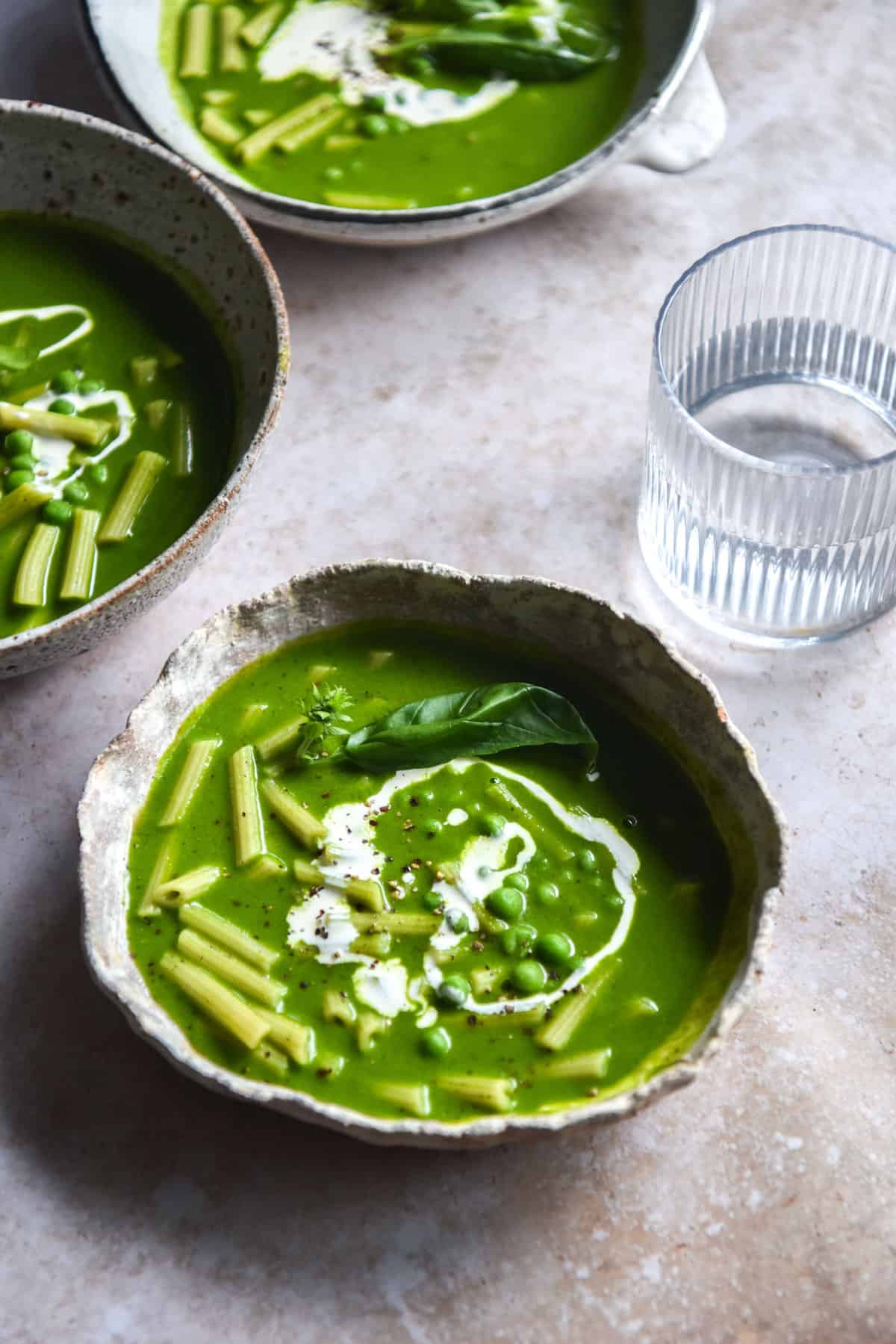 An aerial image of three white ceramic bowls filled with a verdant green pesto pasta soup. The bowls sit atop a beige backdrop and a glass of water sits to the right of the image. 