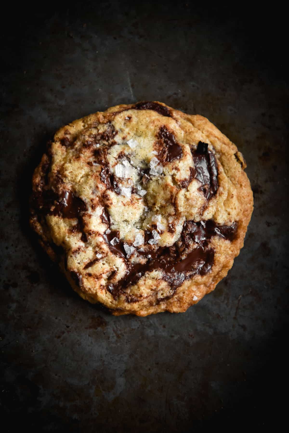 A moody aerial image of a gluten free vegan choc chip cookie on a dark steel backdrop. The cookie is golden brown and dotted with melted chocolate and sea salt flakes. 