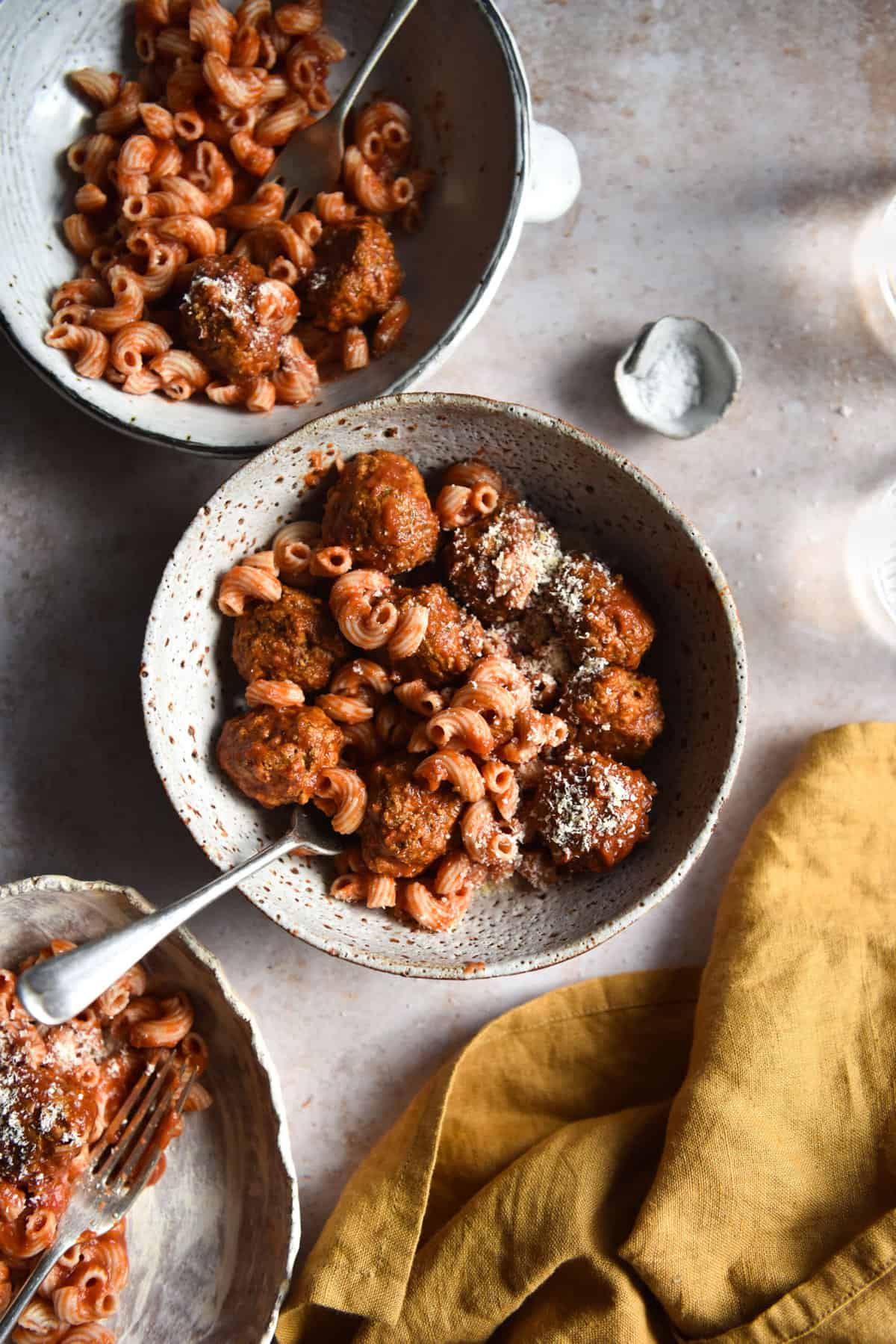 An aerial image of three white ceramic bowls filled with low fodmap vegan meatballs and pasta. The bowls sit atop a light backdrop and are surrounded by glasses of water and a turmeric coloured cloth.