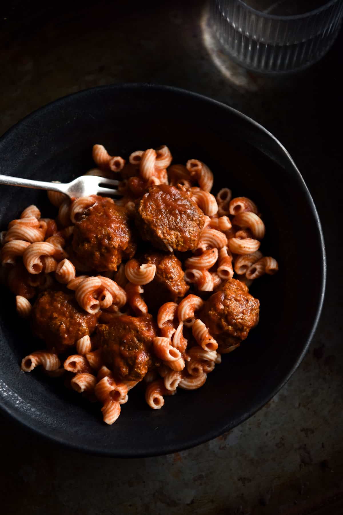 A moody aerial image of a dark ceramic bowl filled with low FODMAP vegan meatballs and pasta. A fork sticks into the bowl from the left hand side and the bowl sits atop a dark steel backdrop.