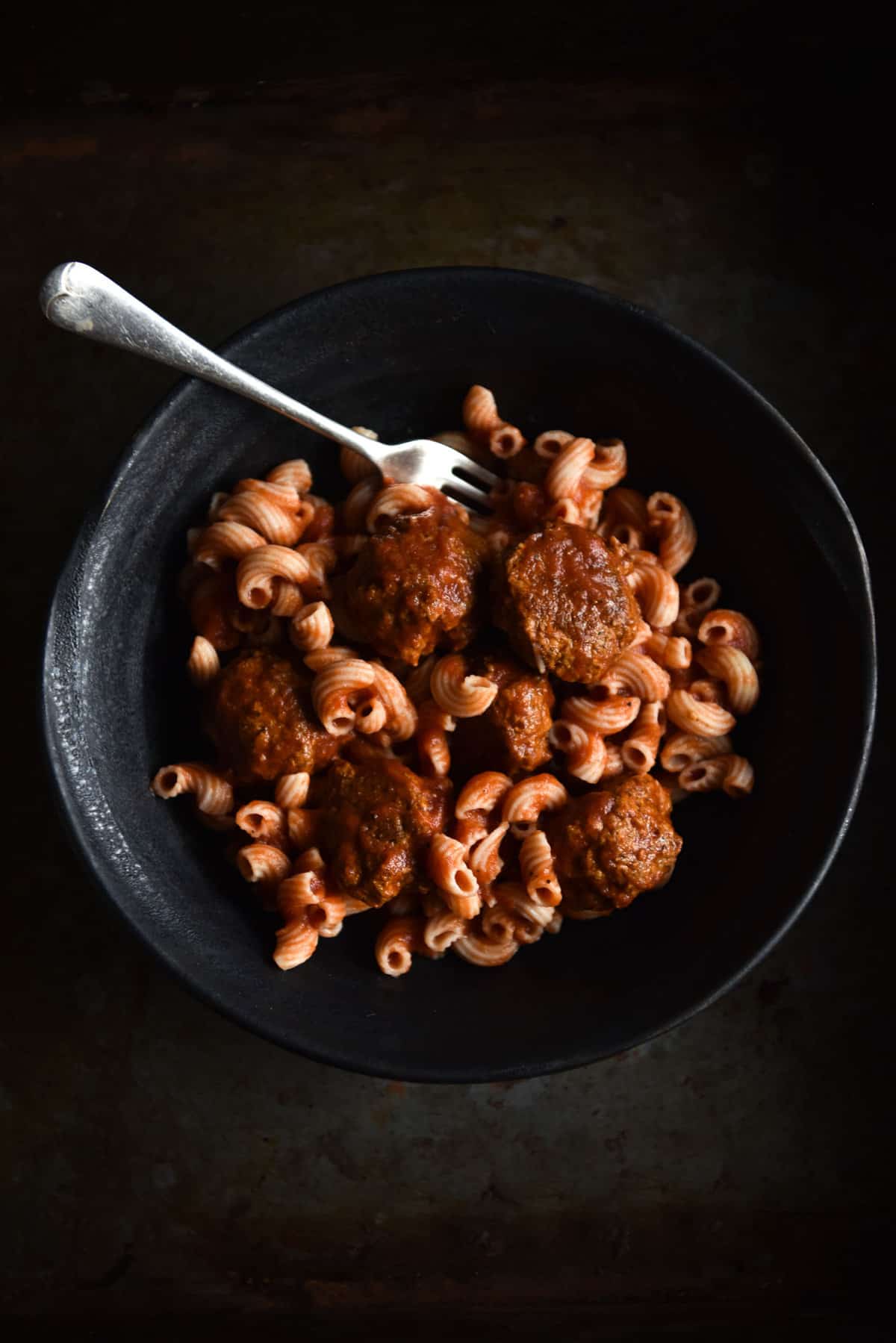 Vegan FODMAP friendly meatballs with a tomato sauce based pasta in a dark ceramic bowl on a dark steel backdrop.