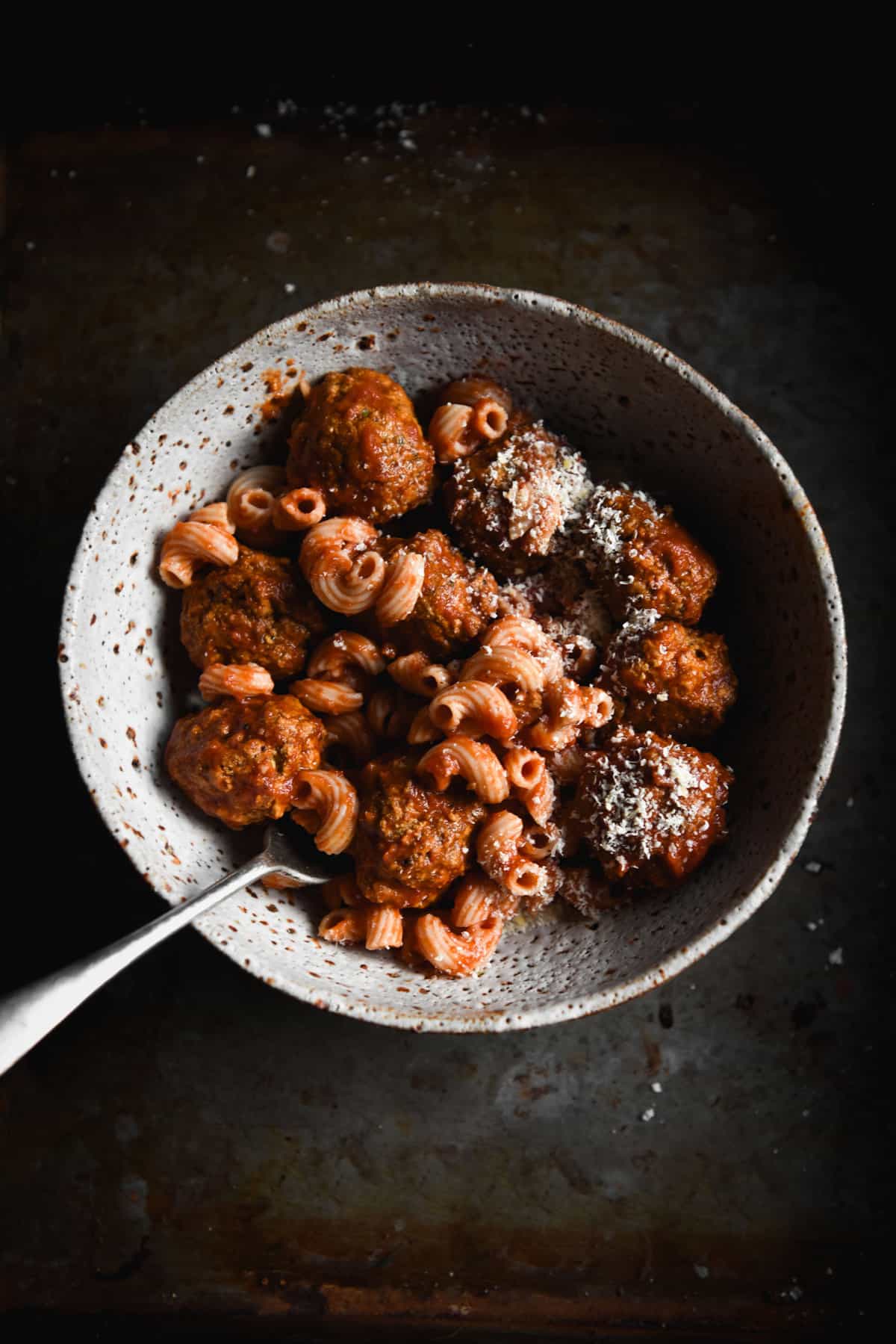 Vegan FODMAP friendly meatballs with a tomato based sauce on gluten free pasta in a white ceramic bowl on a dark steel backdrop. 