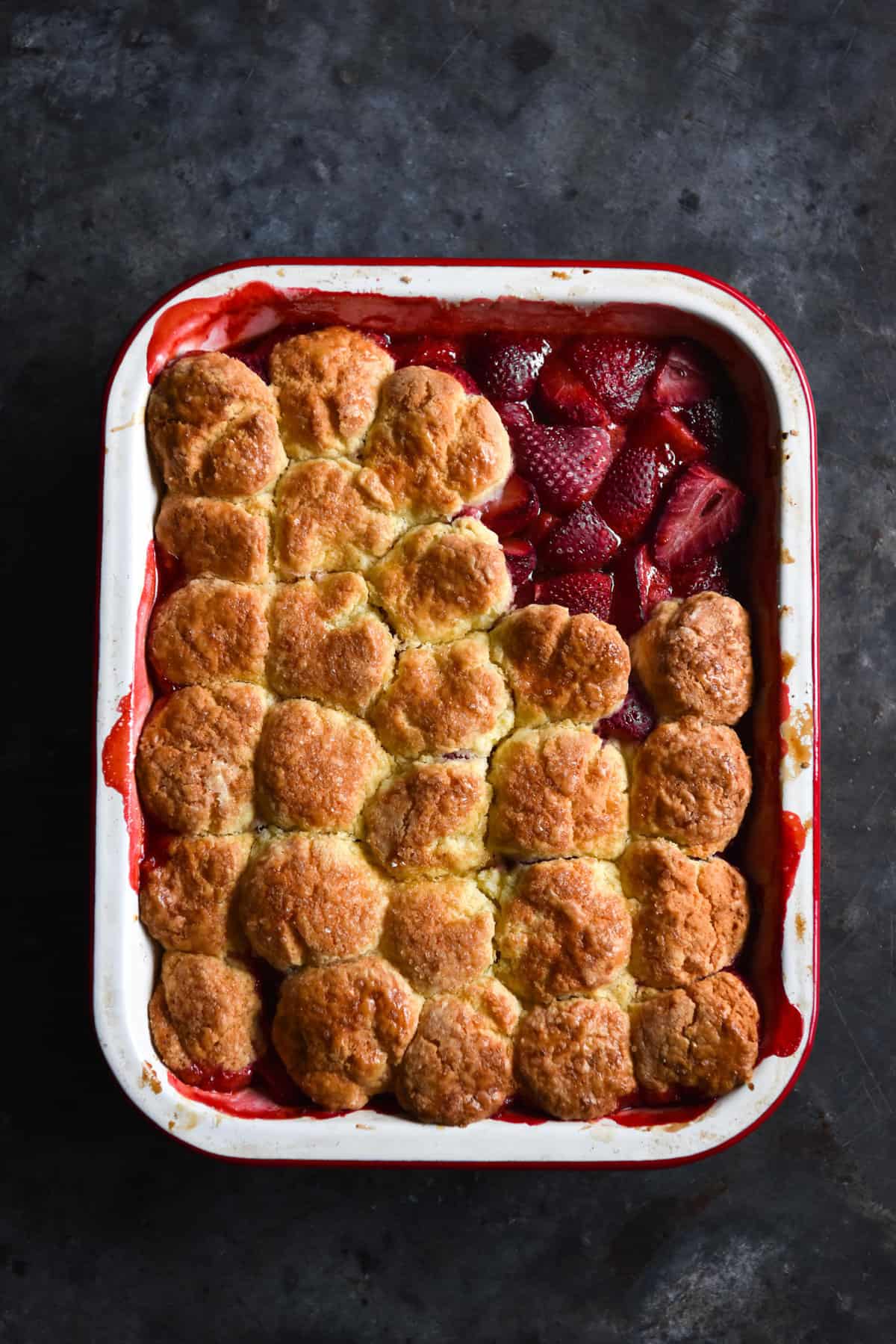 An aerial image of a gluten free cobbler on a dark blue backdrop. The cobbler has cobbler pieces removed from the top right corner, revealing the strawberries underneath.