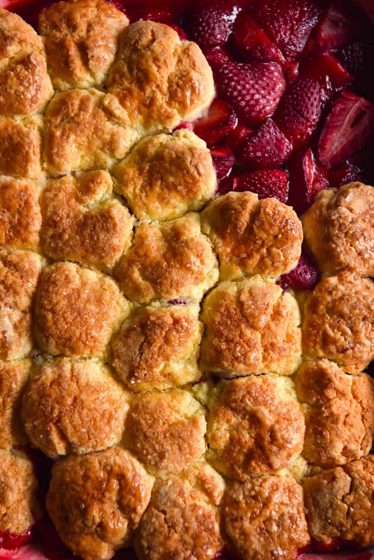 An aerial macro image of a gluten free strawberry cobbler. The cobbler is golden brown and the top right corner of biscuit pieces have been removed, revealing the strawberries underneath.