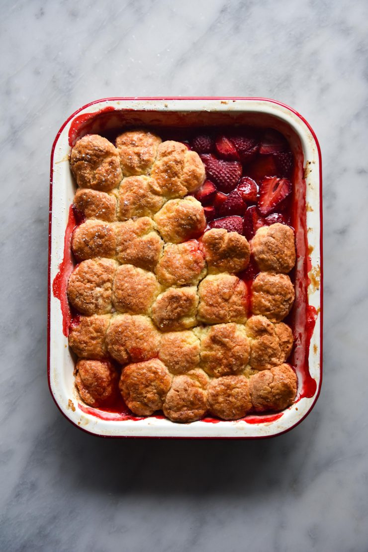 A gluten free strawberry cobbler sits on a white marble table in a rectangular baking dish. Some of the pastry has been taken away, revealing the oozy strawberries underneath.