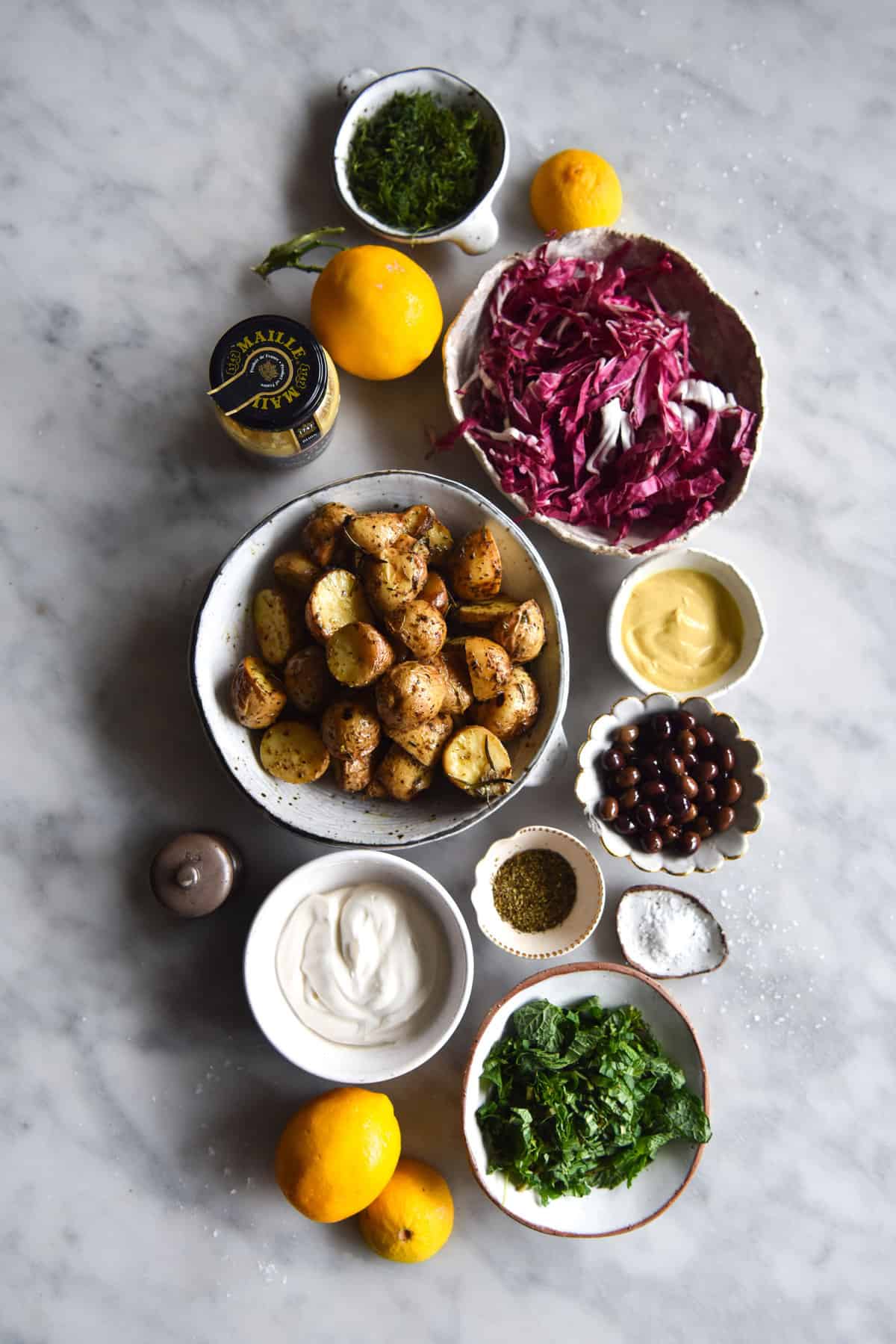 An aerial image of the ingredients used to make a low FODMAP vegan potato salad arranged in small ceramic bowls on a white marble table. 