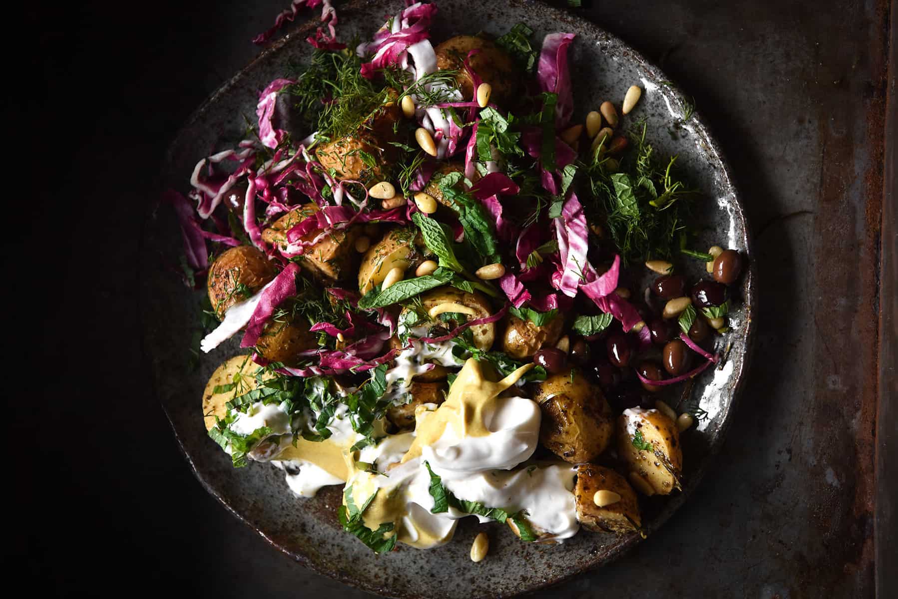 A dark and moody image of a vegan potato salad atop a dark blue ceramic plate on a dark steel backdrop.