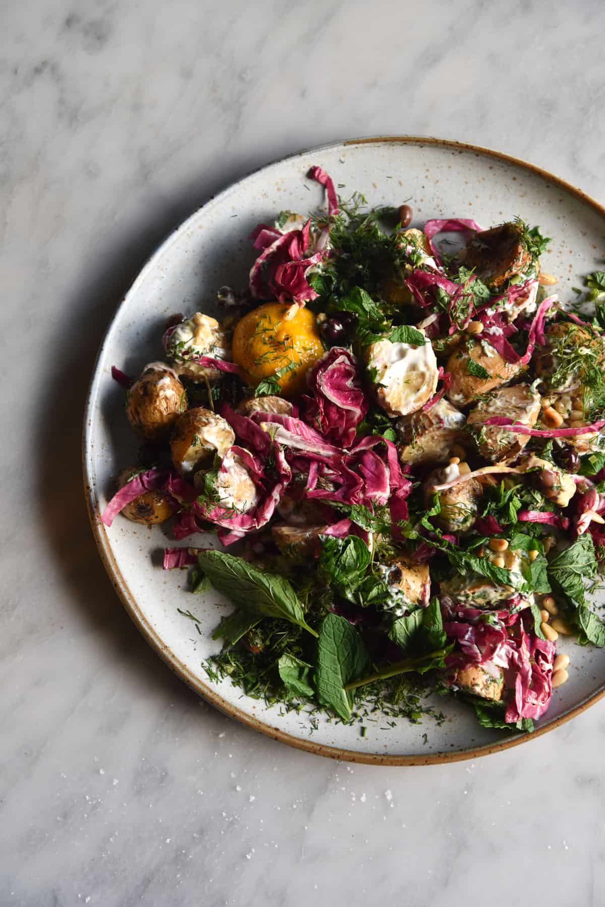 An aerial image of a vegan potato salad with a zingy mayo dressing, radicchio, olives, pine nuts and fresh herbs on a white ceramic plate atop a white marble table