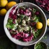 An aerial image of a FODMAP friendly vegan potato salad in a grey ceramic bowl against a moody backdrop. The salad is surrounded by lemons, extra herbs and water glasses. The vibrant radicchio contrasts with the green herbs and mayonnaise coated potatoes
