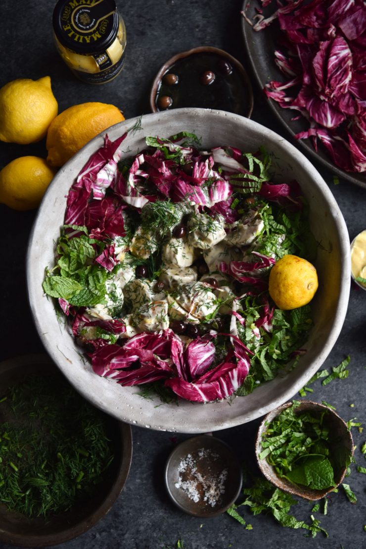 An aerial image of a FODMAP friendly vegan potato salad in a grey ceramic bowl against a moody backdrop. The salad is surrounded by lemons, extra herbs and water glasses. The vibrant radicchio contrasts with the green herbs and mayonnaise coated potatoes