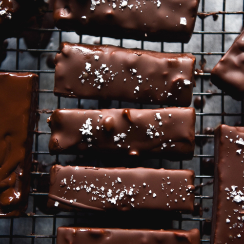 An aerial image of chewy peanut butter bars drying on a baking rack. Their shiny chocolate shells are topped with sea salt flakes and encase toasted peanuts and a peanut centre. They sit atop a cooling rack over a piece of parchment paper.