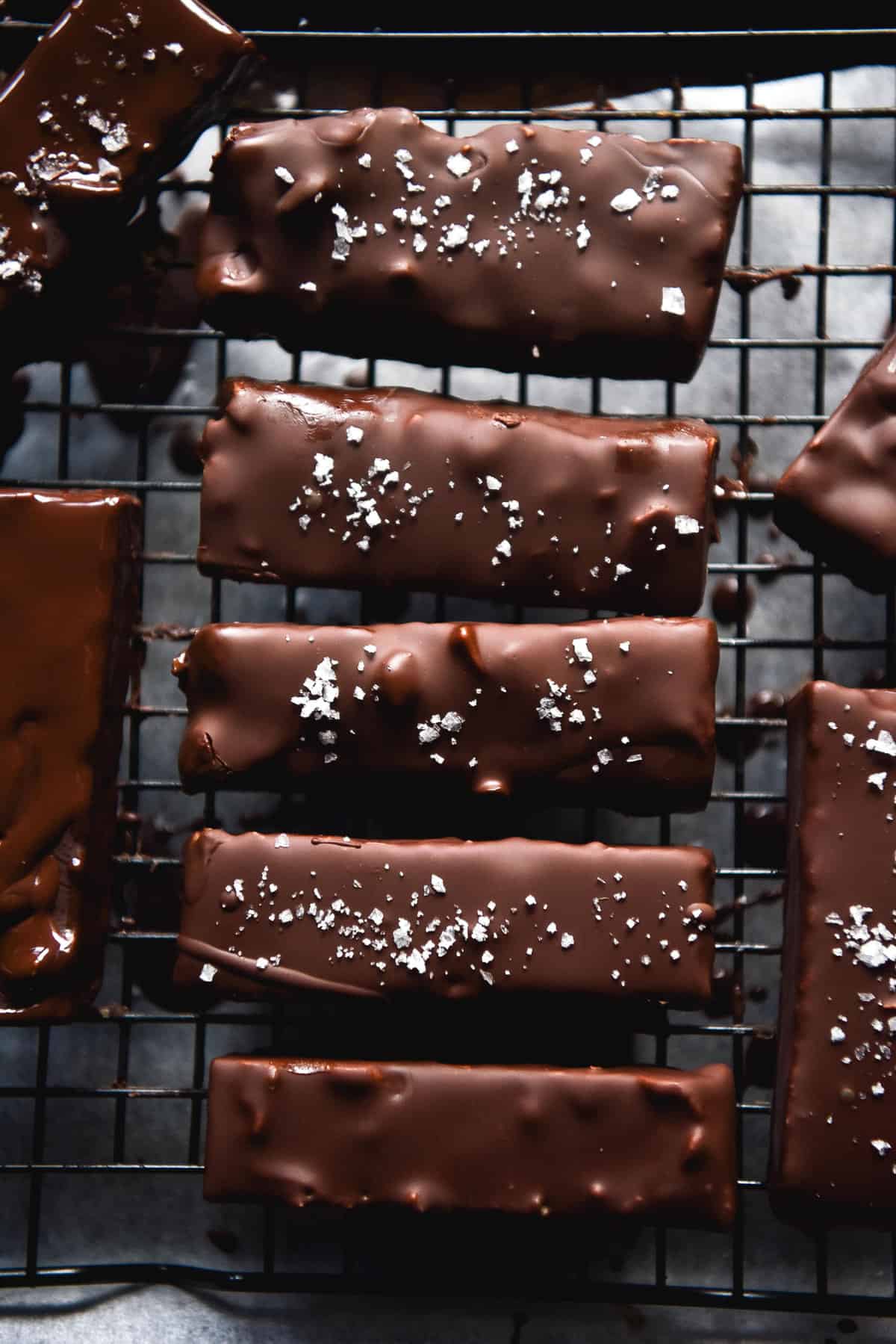 An aerial moody image of chocolate peanut butter bars on a cake rack. The bars are covered in dark chocolate and sprinkled with sea salt flakes.